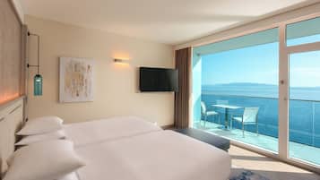 Habitación de hotel con cama, televisor, cuadro y balcón con vista al mar.