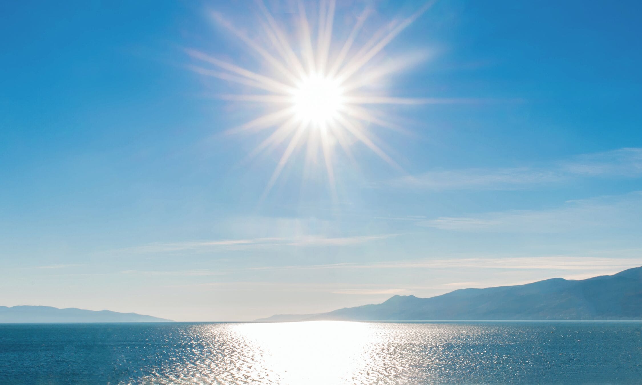 Klarer Himmel mit strahlender Sonne über einem ruhigen Meer und fernen Bergen.