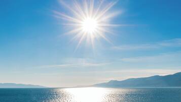 Klarer Himmel mit strahlender Sonne über einem ruhigen Meer und fernen Bergen.