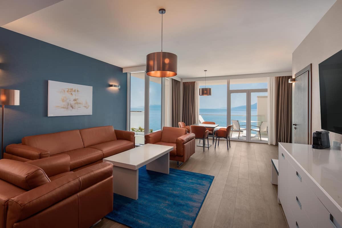 A modern hotel room with a sofa, dining table, and chairs, a large window with a view of the sea, and a flat-screen TV.