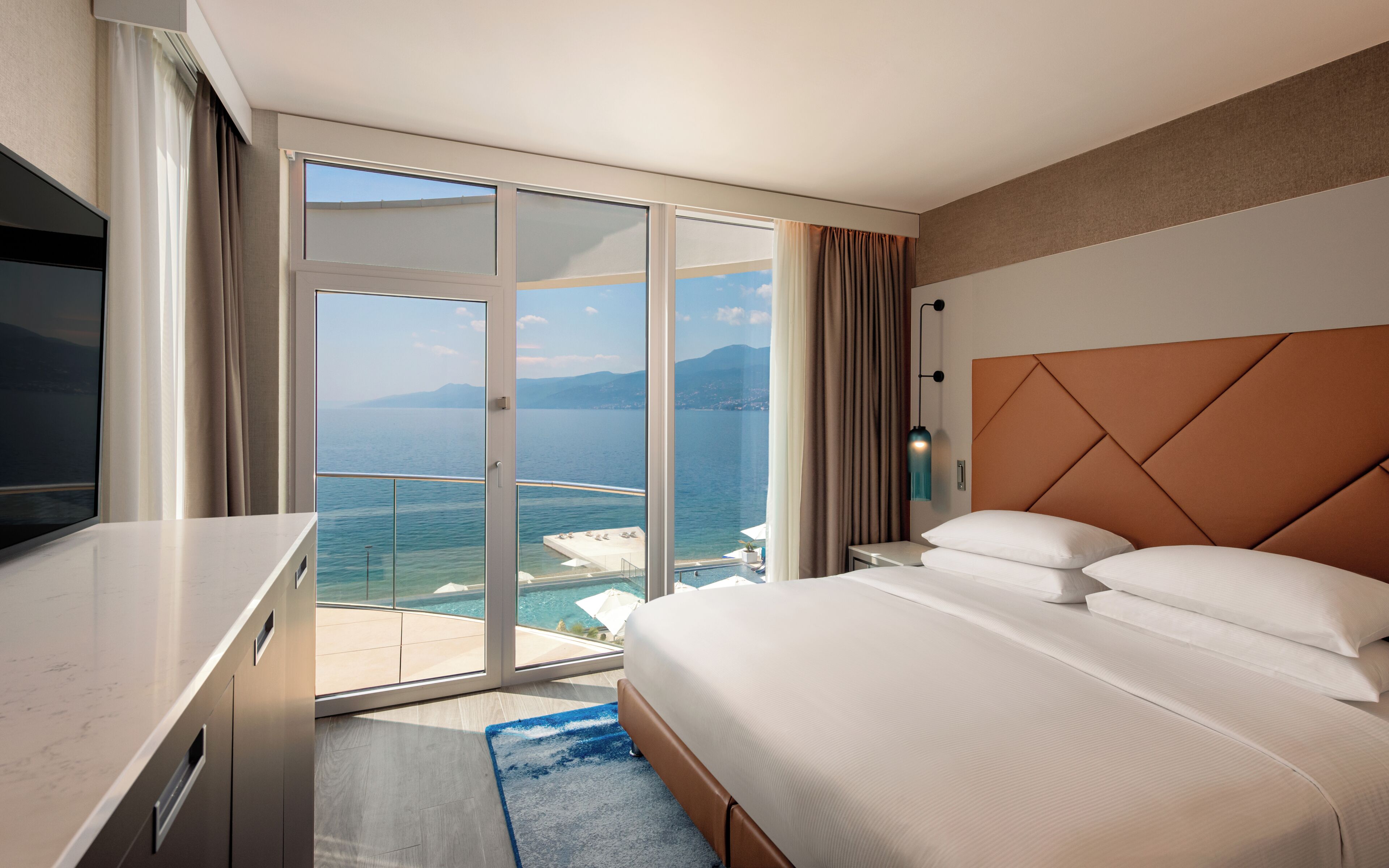 A hotel room with a bed, a TV, a large window with a view of the sea, and a sliding door.