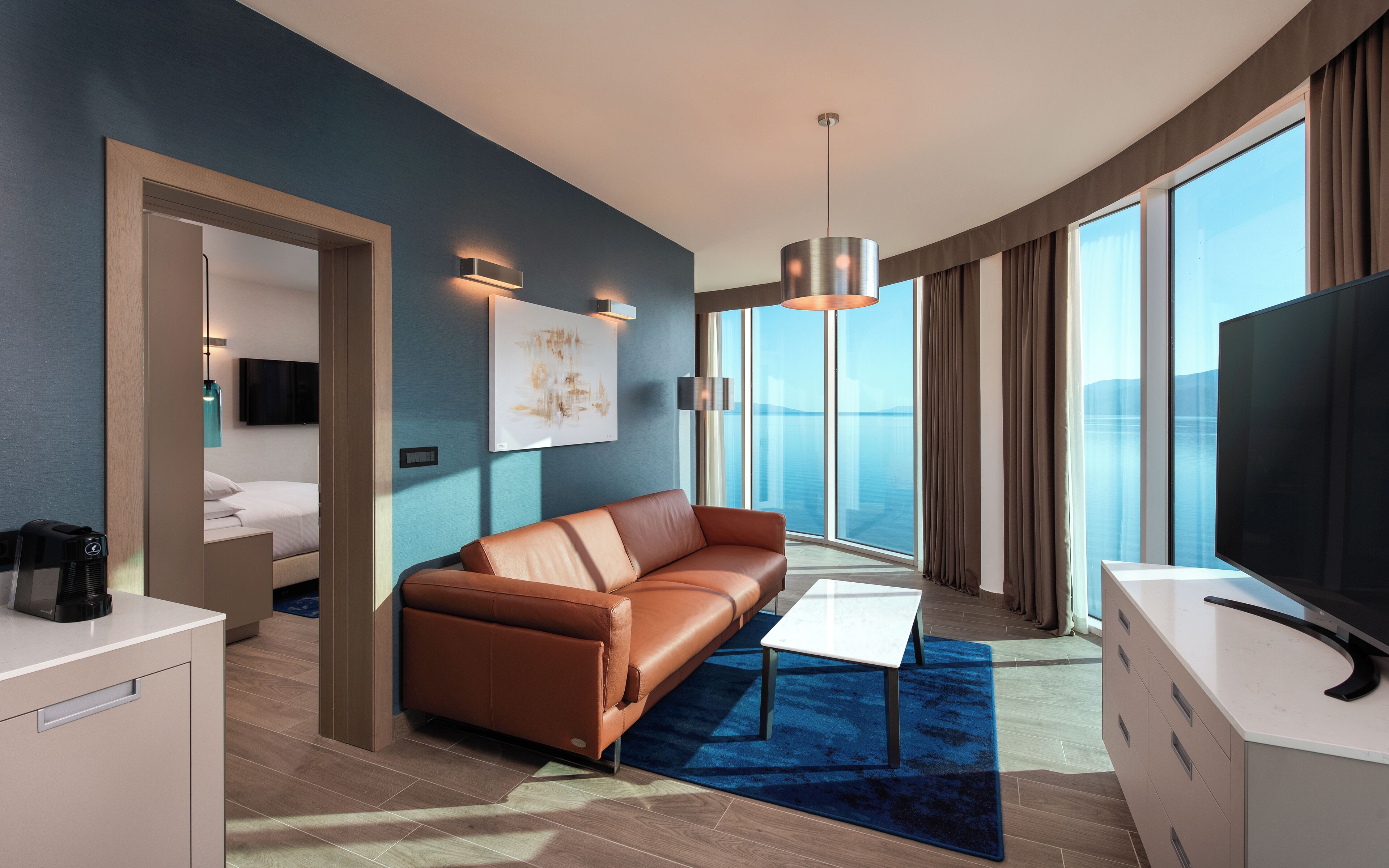 A modern hotel room with a sofa, a coffee maker, a TV, and a large window offering a view of the water.