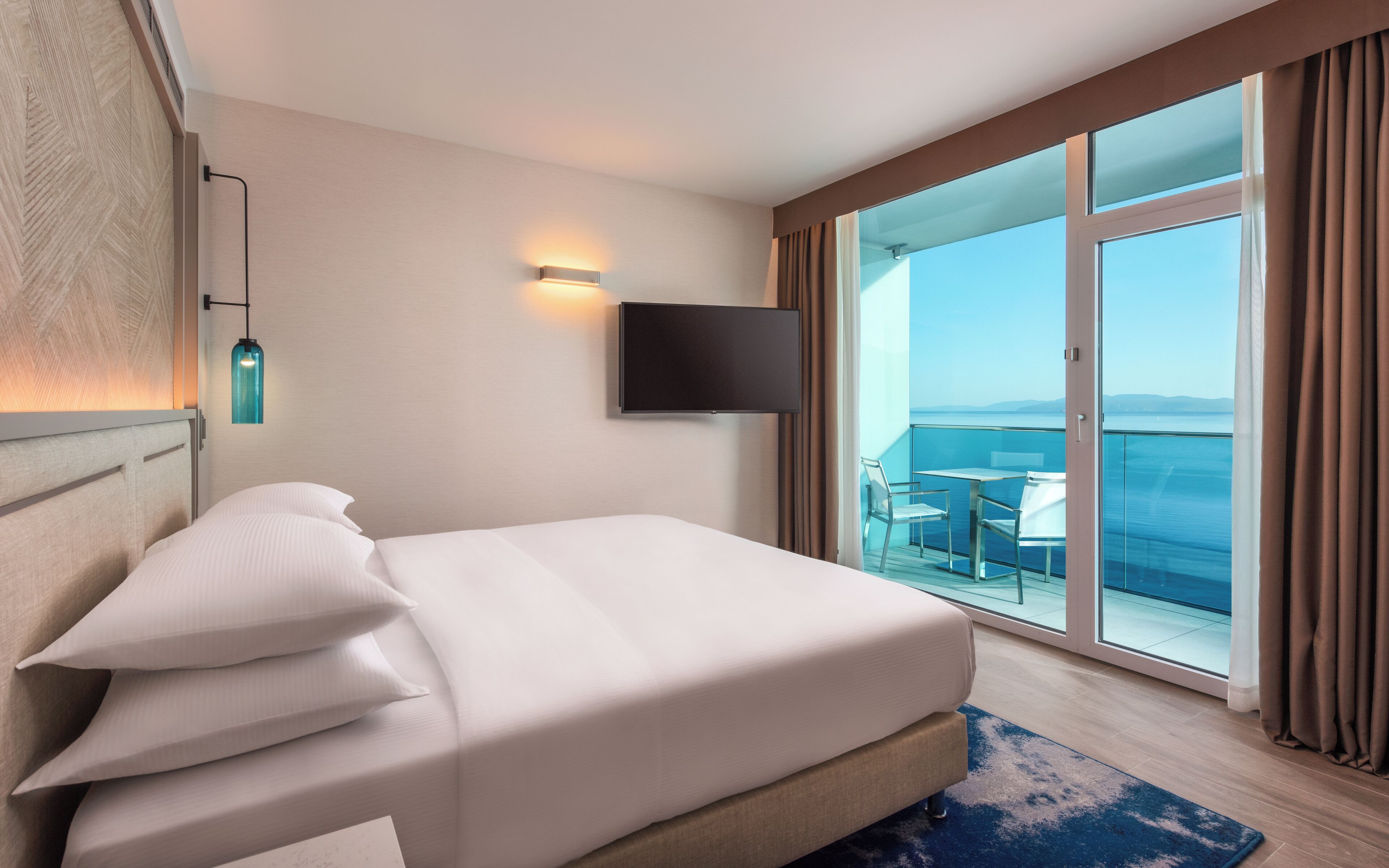 A hotel room with a bed, a TV mounted on the wall, a balcony with a table and chairs, and a view of the sea.