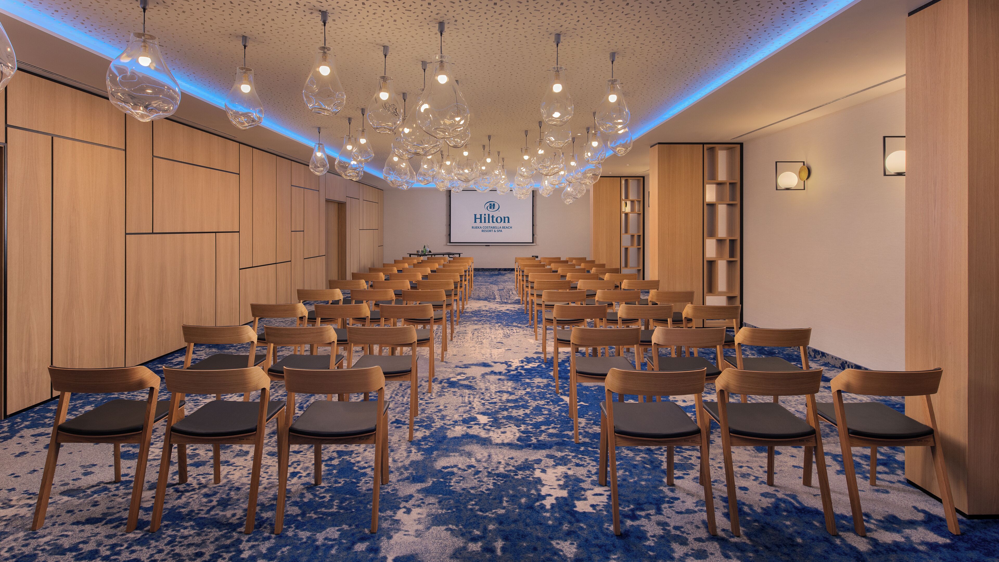 Un salón de conferencias con filas de sillas, una pantalla de proyector y lámparas colgantes.