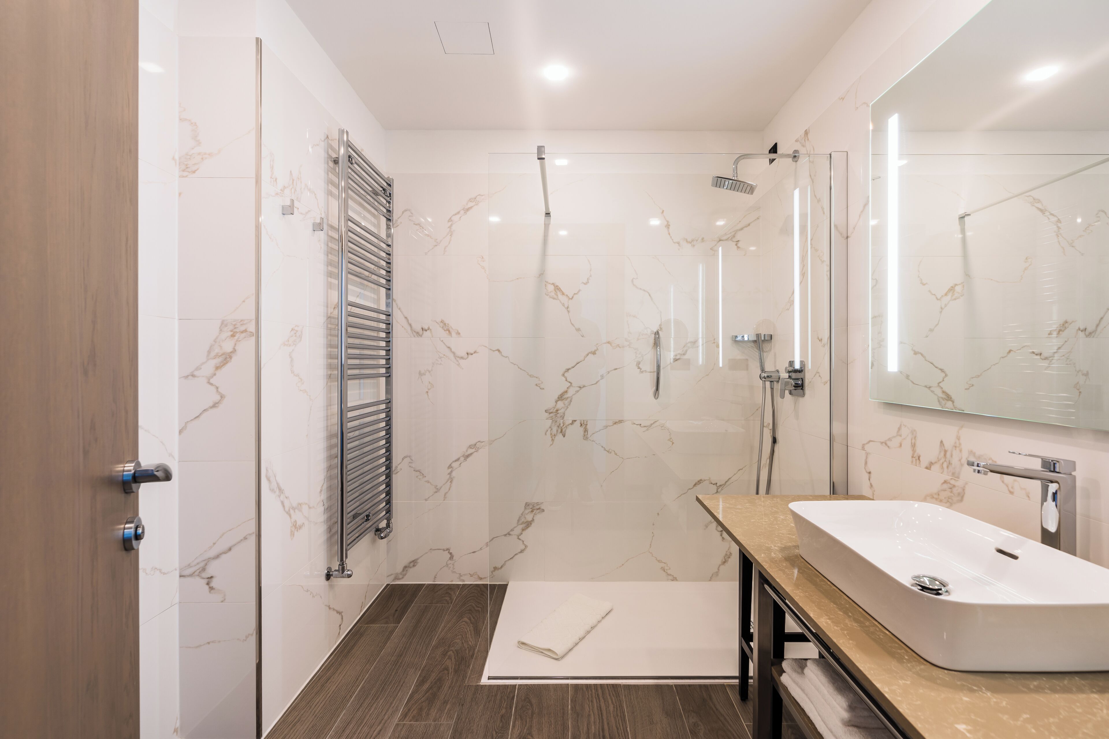 A modern bathroom with a glass shower enclosure, a marble-tiled wall, a wooden floor, and a large mirror above the sink.