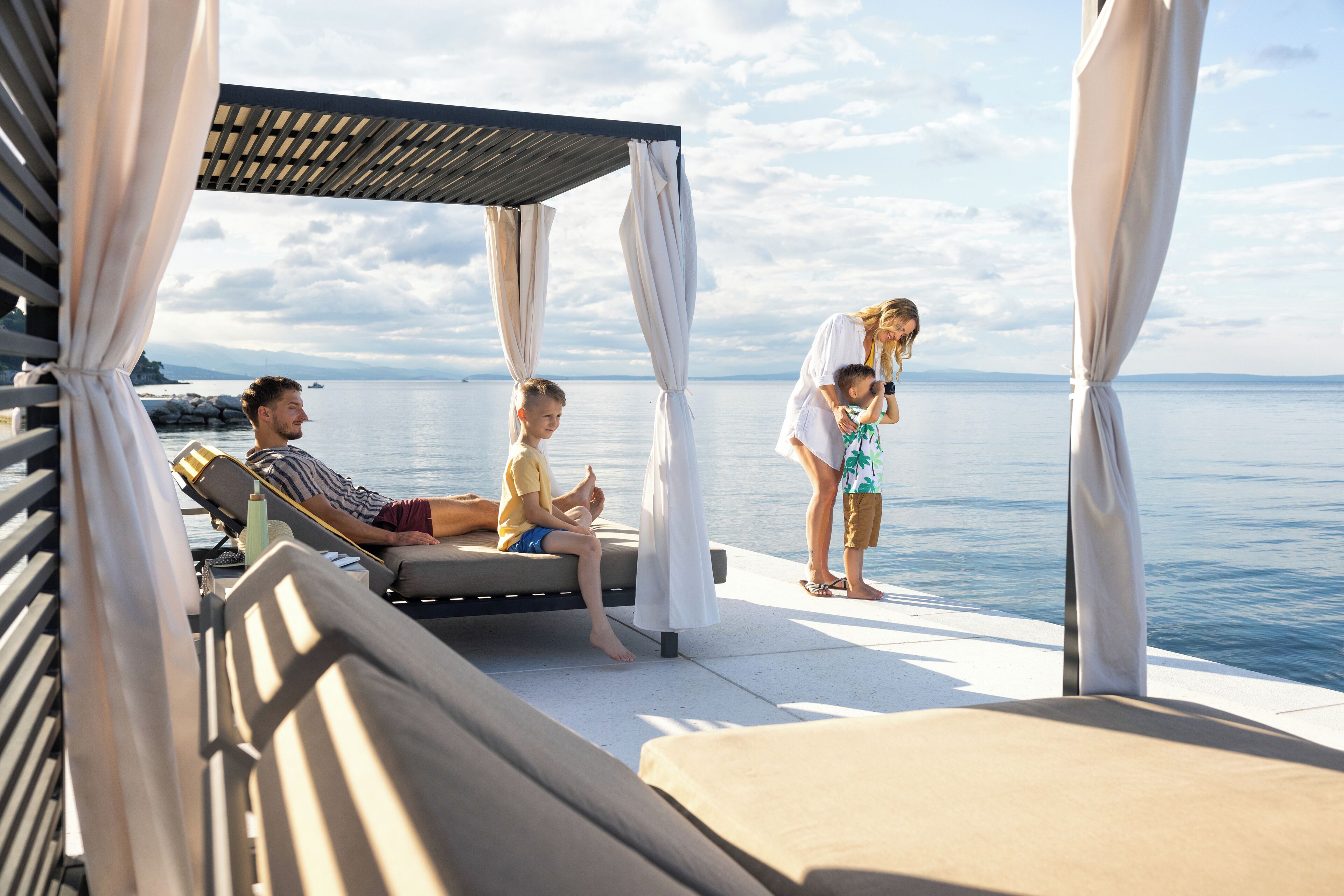 A covered outdoor seating area with a view of the sea, a pool, and a lounge chair.