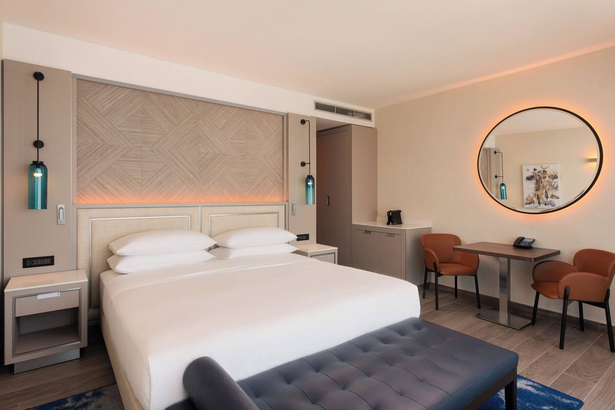 A hotel room with a large bed, bedside tables, a desk with two chairs, a round mirror, and a wall-mounted heater.