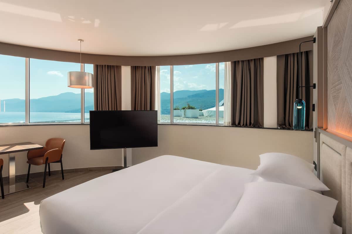A hotel room with a large bed, a TV, a desk with chairs, and a view of the sea.