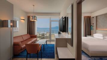 A hotel room with a sofa, a chair, a TV, a bed, and a balcony with a view of the sea.