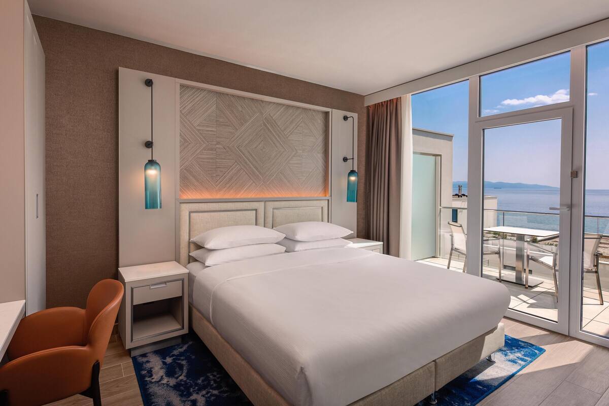A hotel room with a bed, a desk, a chair, a nightstand, and a view of the sea.
