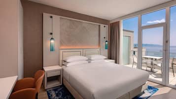 A hotel room with a bed, a desk, a chair, a nightstand, and a view of the sea.