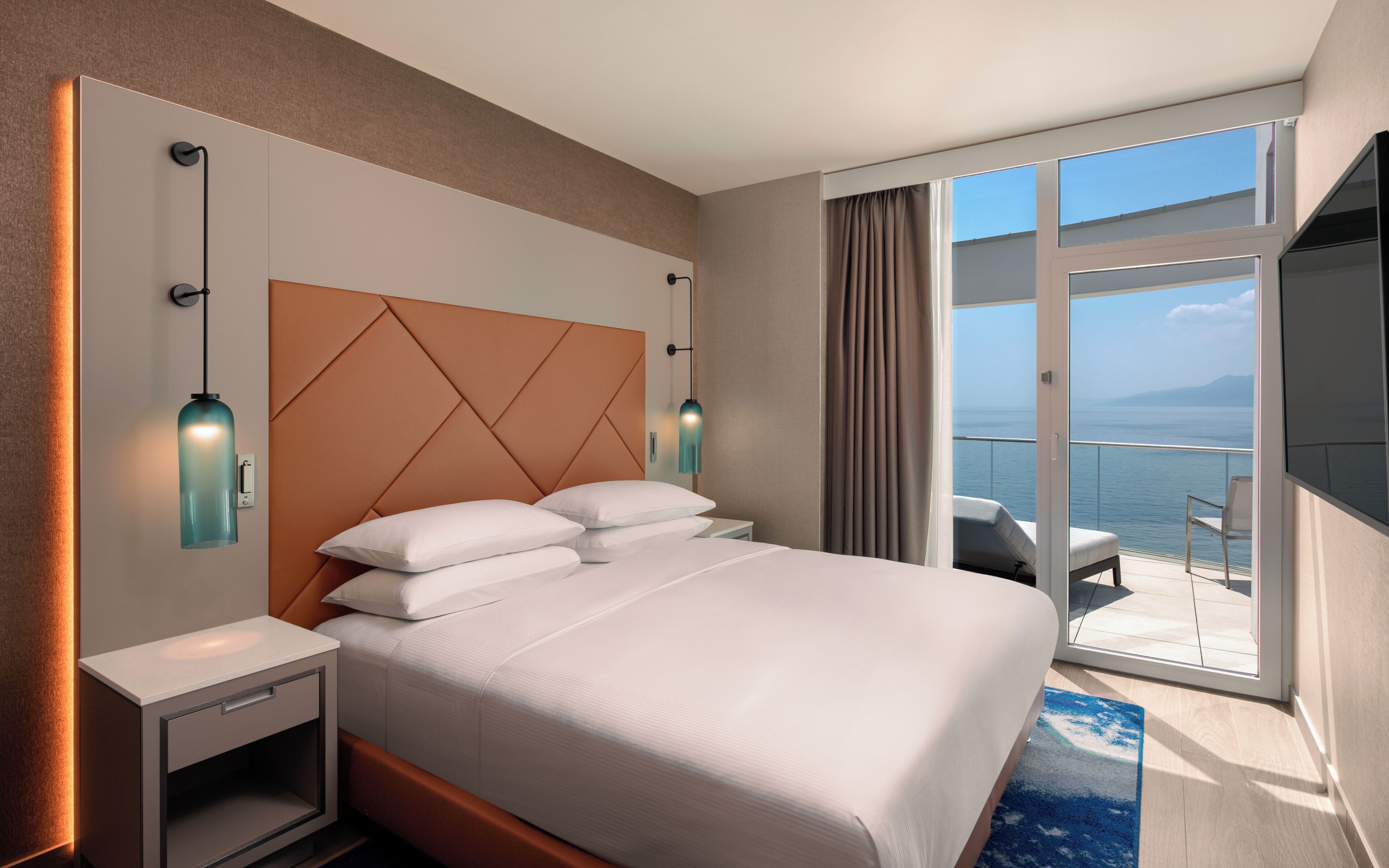 Ein Hotelzimmer mit Bett, Nachttischen, einem Fernseher und einem Balkon mit Meerblick.