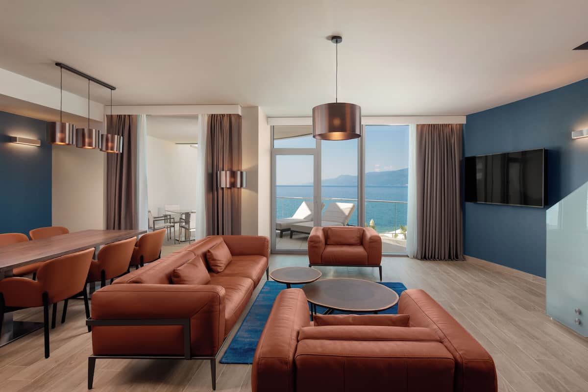 A modern hotel room with a dining area, a sofa set, a TV mounted on the wall, and a balcony with a view of the sea.