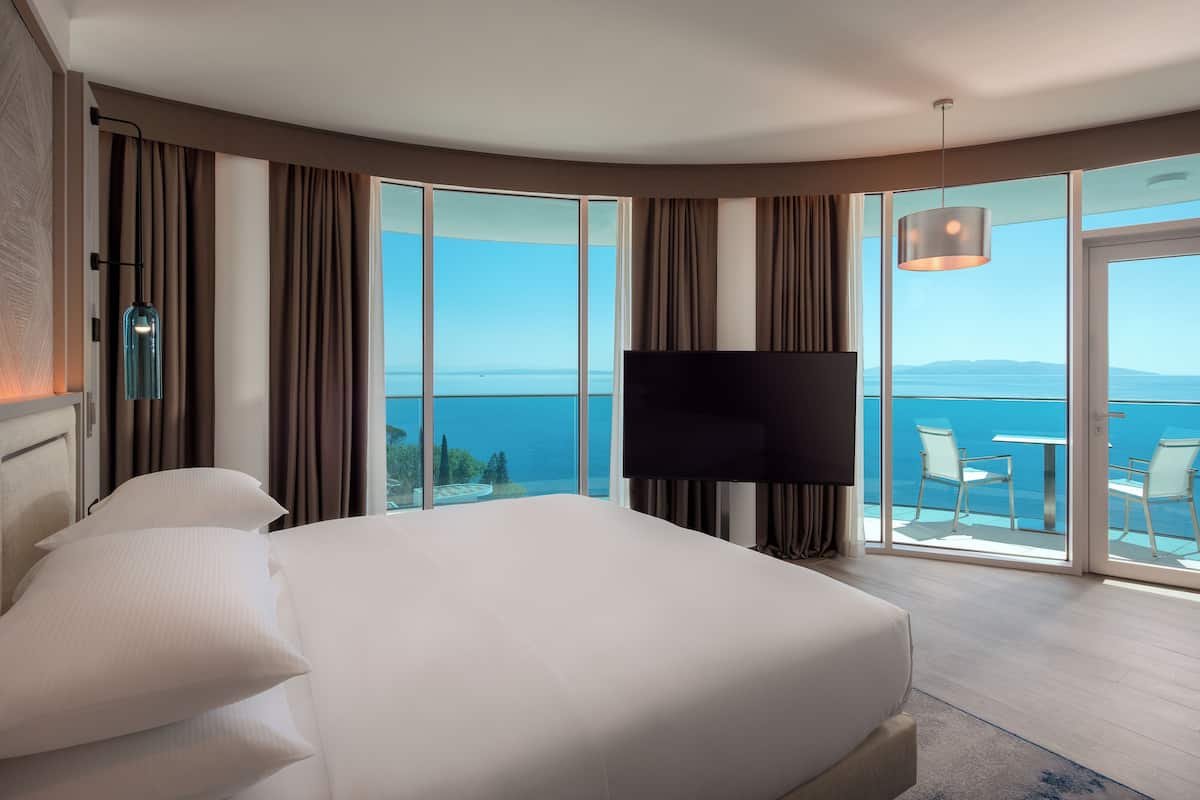 A hotel room with a large bed, a flat-screen TV, a balcony with a table and chairs, and a view of the sea.