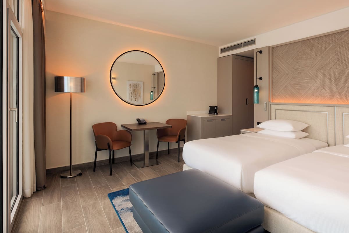 A hotel room with two beds, a desk, chairs, a round mirror, and a small table.
