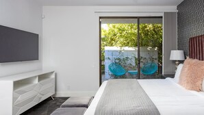 Une chambre moderne dotée d’un téléviseur à écran plat, d’un lit recouvert d’une courtepointe matelassée, d’une table de chevet avec une lampe, et offrant une vue sur un balcon agrémenté de chaises et de plantes.