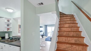 Una cocina con pileta, lavavajillas y muebles bajos blancos. Una escalera con escalones de madera lleva a un balcón.