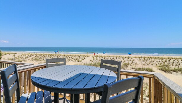 Una terraza de madera con una mesa redonda y sillas, con vista a una playa, dunas y el océano.