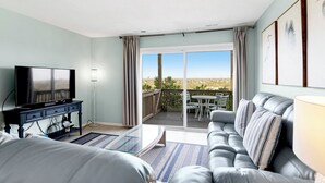 Uma sala de estar com televisão, sofá e varanda com mesa e cadeiras.