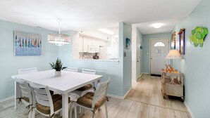 Uma sala de jantar com uma mesa e cadeiras brancas, um lustre e vista para a cozinha.