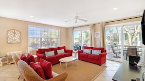 Ein geräumiges Wohnzimmer mit zwei roten Sofas, einem Korbstuhl, einem Couchtisch und einem Deckenventilator. Große Fenster sorgen für viel natürliches Licht und bieten einen Blick ins Freie.
