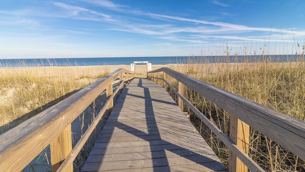 Ein hölzerner Steg führt zu einem Strand unter blauem Himmel.