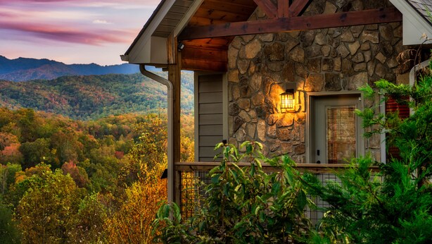 Una casa de piedra con porche de madera, chimenea de piedra y vista a una cordillera boscosa.