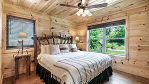 Un dormitorio de cabaña de madera con una cama, dos lámparas, un ventilador de techo y una ventana con vistas a la vegetación.