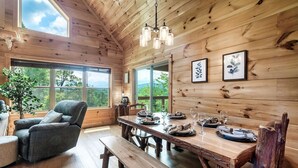 Interior de una cabaña de madera con una mesa de comedor dispuesta para cuatro personas, un sillón reclinable y grandes ventanales que ofrecen vistas al exterior.
