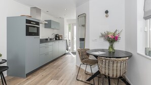 Et moderne køkken med lysegrå skabe, en sort bordplade og en indbygget ovn. Der er et spiseområde med et rundt bord og fletstole. Et spejl hænger på væggen, og en vase med blomster står på bordet.