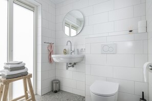 A modern bathroom with white tiles, a round mirror, a sink, a toilet, and a small trash bin. - For a wonderful vacation for two by the sea near Domburg (Aagtekerke)
