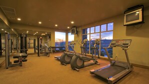 A gym with treadmills, elliptical machines, and weightlifting equipment, with a large window offering a view of the cityscape. - Zions Gateway Escape (Hurricane)