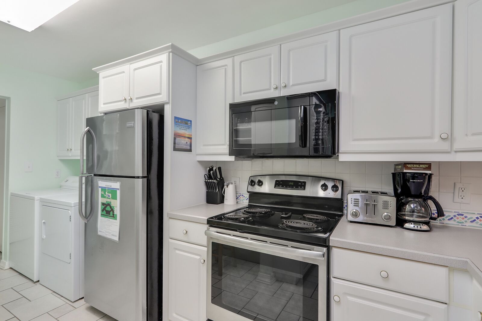 A modern kitchen with white cabinets, a stainless steel refrigerator, a microwave, a stove, and a coffee maker.