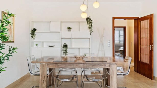 Zona pranzo con tavolo in legno e sedie trasparenti, lampadario a sospensione e mobiletto bianco con piante.