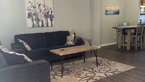 Una sala de estar con un sofá de color oscuro, una mesa de centro de madera, un tapete estampado, un ventilador de techo y obras de arte en las paredes.