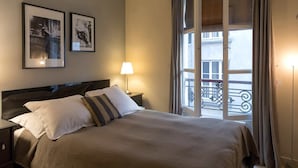 A hotel room with a bed, a nightstand, a lamp, and two framed pictures on the wall. - Rivoli Market I Comfortable 1 Bed Apt near Hotel de Ville (Paris)