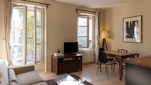A living room with a sofa, a coffee table, a TV on a wooden cabinet, a dining table with chairs, and a lamp. - Rivoli Market I Comfortable 1 Bed Apt near Hotel de Ville (Paris)