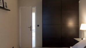 A bedroom with a bed, a nightstand with a lamp, a dark wardrobe, and a white door leading to a bright room. - Rivoli Market I Comfortable 1 Bed Apt near Hotel de Ville (Paris)
