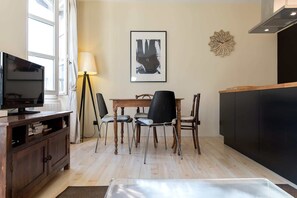 A compact living space with a wooden dining table, four chairs, a television on a wooden stand, a floor lamp, and a framed picture on the wall.