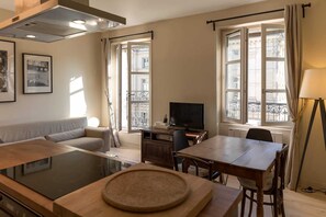 A modern living room with a sofa, a dining table with chairs, a TV, and a kitchen area with a stove. - Rivoli Market I Comfortable 1 Bed Apt near Hotel de Ville (Paris)