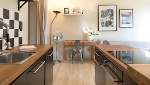 A modern kitchen with a central island, a dining area with a chair, and a sofa. There are framed pictures on the wall and a floor lamp. - Rivoli Market I Comfortable 1 Bed Apt near Hotel de Ville (Paris)