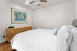 Une chambre à coucher avec un lit, une commode en bois, un ventilateur de plafond et un tableau encadré accroché au mur.