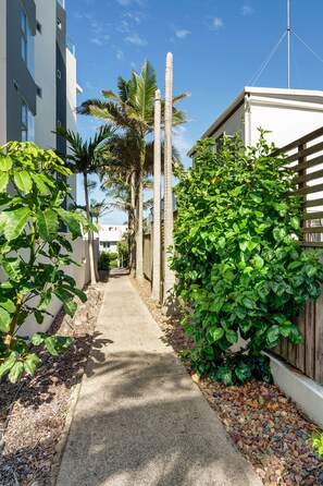 Un étroit sentier en béton, bordé d’une végétation luxuriante et d’une clôture en bois, mène à un bâtiment blanc doté d’un balcon.