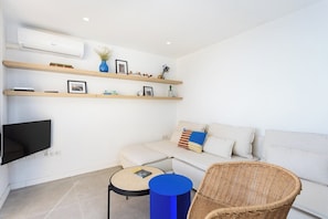 Una sala de estar moderna con televisor de pantalla plana, un sofá, una silla de mimbre, una mesa de centro redonda y estantes empotrados con objetos decorativos.