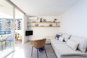 A modern living room with a sofa, a wicker chair, a TV mounted on the wall, and a balcony with a view of buildings.
