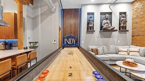 Ein Shuffleboard-Tisch mit nummerierten Enden, eine Bar mit Hockern und eine Sitzecke mit einer Couch und einem Couchtisch.