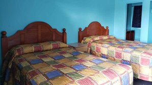 A room with two wooden beds with colorful quilts, a small table, and a door leading to another room.