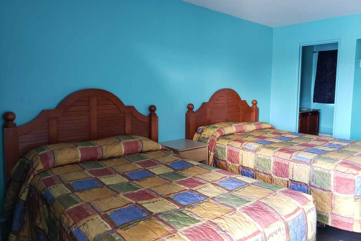 Quarto com duas camas de madeira, cobertores coloridos, uma pequena mesa e uma porta que dá para outro cômodo.