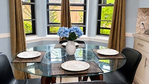 A dining area with a glass table surrounded by chairs, a vase with flowers, and a view of greenery through large windows. - Family Ready Spacious Retreat - 4BR, Sleeps 8, Pet Friendly with Gazebo (Houston)