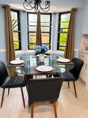 Un coin repas avec une table en verre entourĂ©e de chaises, un vase rempli de fleurs et une vue sur la verdure grĂące Ă de grandes fenĂȘtres.